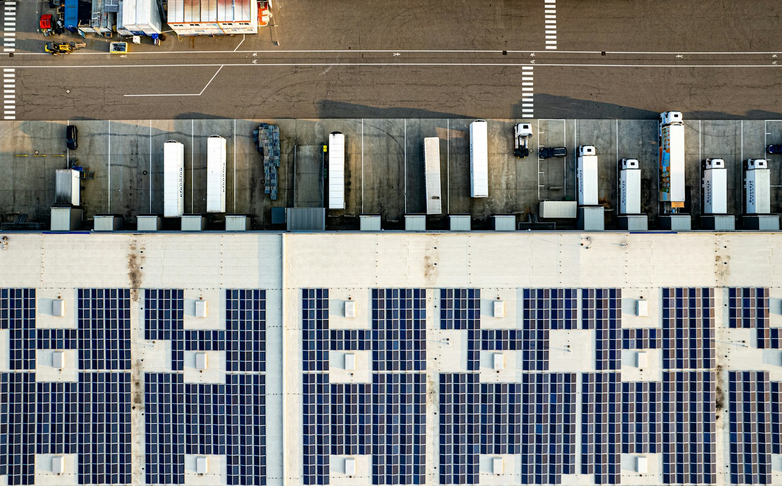 Letecký pohled na střechu logistického skladu pokrytou fotovoltaickými panely, před budovou stojí nákladní vozy u nakládacích ramp.